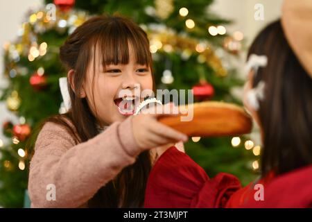 Adorabili sorelline in caldi abiti invernali che mangiano pane fresco vicino all'albero di Natale decorato Foto Stock