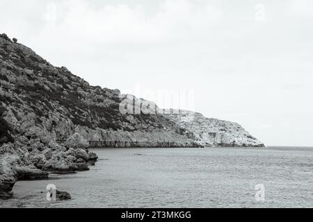 Vista in bianco e nero della collina di formazione rocciosa sulla spiaggia di Anthony Quinn Bay nell'acqua dell'oceano Foto Stock
