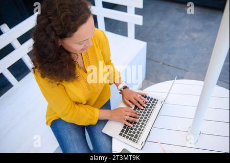 Vista dall'alto di un'affascinante bruna dai capelli ricci multietnici, imprenditrice, freelance che lavora a distanza su un computer portatile, seduta su un legno bianco Foto Stock