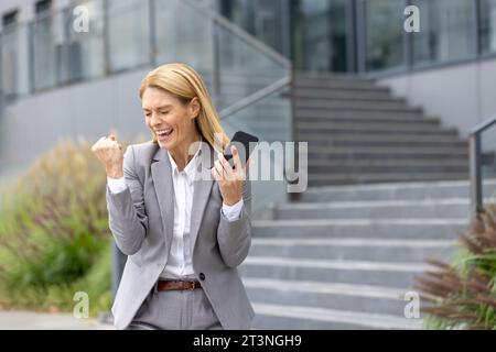 Una donna d'affari matura e gioiosa di successo ha ricevuto un messaggio di notifica online con buoni risultati al telefono, il capo donna utilizza l'app sullo smartphone, cammina fuori dall'edificio dell'ufficio in tuta aziendale. Foto Stock