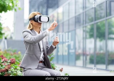 Donna d'affari fuori dall'edificio degli uffici utilizza occhiali per realtà virtuale, boss donna siede su una panchina di giorno in una tuta da lavoro. Foto Stock