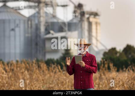 Agricoltore anziano soddisfatto con un cappello di paglia che guarda una tavoletta davanti ai silos di grano nel campo di mais durante il raccolto Foto Stock