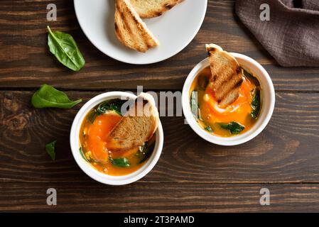 Uova cotte al forno (uova e cocotte) con crema, сheese e spinaci su fondo di legno. Servito con pane tostato caldo per condire. Vista dall'alto, base piatta Foto Stock