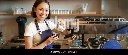 Ragazza barista asiatica sorridente, preparare caffè, versare latte al vapore nel cappuccino, fare latte art in tazza, lavorare in caffè Foto Stock