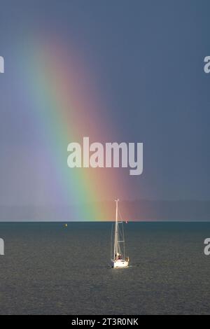 uno yacht solitario che naviga sul mare o sull'oceano alla fine di un arcobaleno attraverso il cielo. barca a vela sotto un impressionante arcobaleno in una giornata tempestosa in mare. Foto Stock