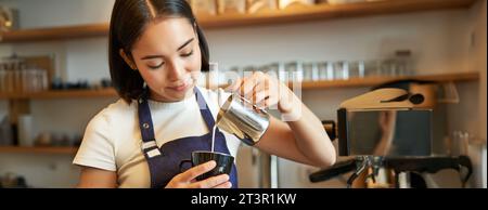 Ragazza barista asiatica sorridente, preparare caffè, versare latte al vapore nel cappuccino, fare latte art in tazza, lavorare in caffè Foto Stock