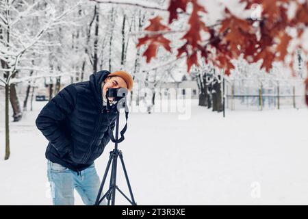 Fotografo che scatta foto utilizzando una fotocamera digitale professionale su cavalletto in un parco invernale innevato guardando nel mirino. Immagini di paesaggio. Fotogr. Per esterni Foto Stock