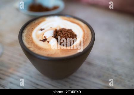 Zucchero granulato di canna scura prima di mescolare sulla schiuma di un caffè bianco piatto su un tavolo di legno in una tazza senza manico. Foto Stock