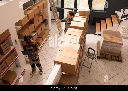 Vista dall'alto di diversi colleghi che lavorano in magazzino, preparando gli ordini dei clienti prima di iniziare a spedire le merci. Personale del magazzino in tuta protettiva che lavora con scatole di cartone Foto Stock