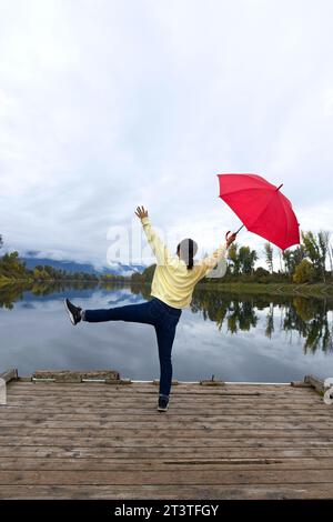 Una donna che tiene un ombrello rosso si mette in posa mentre si trova su un molo vicino al fiume Kootenai nell'Idaho settentrionale. Foto Stock