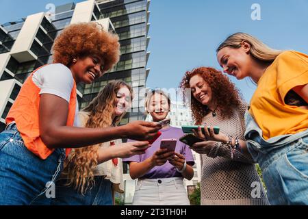 Gruppo di giovani donne felici che guardano i loro smartphone dipendenti dall'app social media e si divertono insieme all'esterno. Donne multirazziali che usano i cellulari e si divertono a giocare a divertenti videogame online. Foto di alta qualità Foto Stock