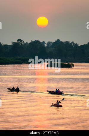 Sagome di figure umane in piccole imbarcazioni strette, che si spostano attraverso le calme e tranquille acque del Mekong, attraverso raggi di luce dorata riflessa su di esso Foto Stock