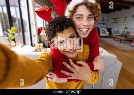 Ritratto di una coppia gay felice e diversificata che indossa cappelli di natale e che fa videochiamata a casa Foto Stock