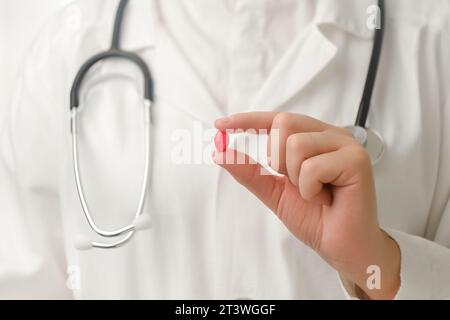 Primo piano di una giovane medico femminile irriconoscibile in uniforme bianca con stetoscopio che tiene in mano una piccola vitamina rossa. In trattamento con vitamine o mediche Foto Stock