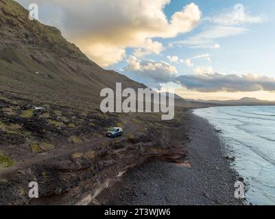 Prospettiva aerea di un SUV turistico che passa su una strada sterrata lungo la costa di Lanzarote e alte scogliere ricoperte di vegetazione con nuvole mattutine Foto Stock