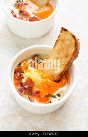 Uova in cocotte (uova cotte al forno) con panna, сheese e pancetta in un recipiente in ceramica su sfondo di pietra chiaro. Servito con pane tostato caldo per condire. Foto Stock