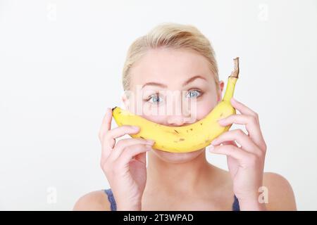 Giovane donna bionda modello caucasico su sfondo bianco tenendo sana banana come parte di un sorriso viso - mangiare sano concetto Foto Stock