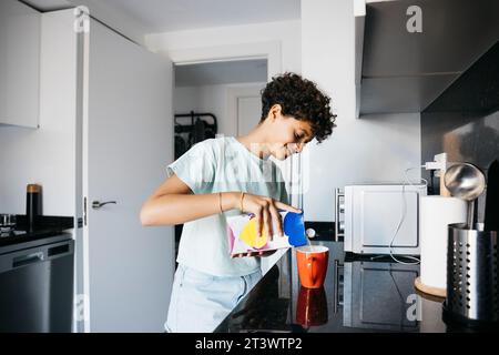 Una bambina di dieci anni che versa latte su una tazza rossa in cucina. Ragazza a casa che prepara una tazza di latte. Foto Stock