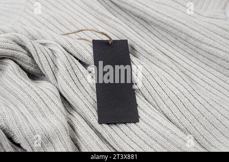Etichetta di cartone nera vuota con cordicella sul maglione grigio. Etichettare il mockup vuoto su un abito, per posizionare il tuo disegno. Prezzo, vendita, indirizzo etichetta. Perno di articolazione Foto Stock