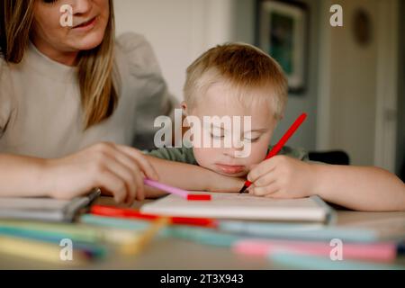 Ragazzo con sindrome di Down che disegna in libro da madre a casa Foto Stock