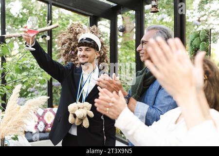 Giovane che solleva brindisi mentre sta con la famiglia applaudendo durante la festa Foto Stock