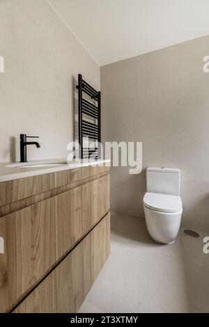 Immagine del bagno con piastrelle color crema, piano di lavoro coordinato, armadio in legno con cassetti e rubinetti neri coordinati con radiatore per asciugamani Foto Stock