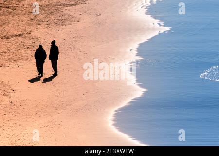 Due persone viste in silhouette che camminano lungo il litorale di Fistral Beach a Newquay, in Cornovaglia, nel Regno Unito. Foto Stock