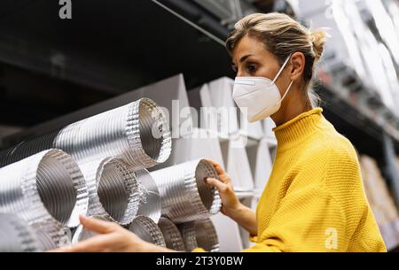 la donna che indossa una maschera facciale e un maglione giallo esamina i condotti di ventilazione in argento in un ambiente di magazzino o di negozio. Immagine del concetto di sicurezza corona Foto Stock