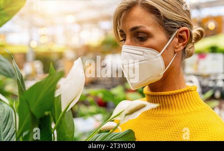 donna con un maglione giallo che indossa una maschera facciale bianca si erge in mezzo al verde lussureggiante, apprezzando un fiore bianco, con uno sfondo del negozio leggermente illuminato. Shoppi Foto Stock