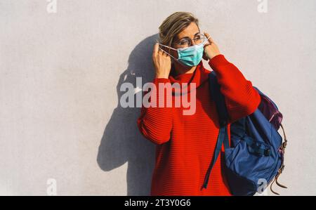 donna con occhiali rotondi si toglie la maschera viso turchese dal naso, vestita con un vivace maglione rosso, con un'ombra distintiva dietro di lei su un contropiede Foto Stock