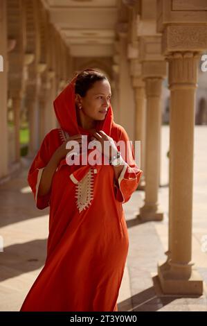 Bella giovane donna bruna mediorientale con abito etnico arancione e testa ricoperta di hijab, in posa sullo sfondo della moschea, in piedi Foto Stock