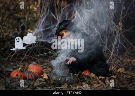 Buon Halloween. il piccolo stregone eroga pozione nel calderone della strega, l'incantesimo del casting. Ragazzo che si diverte, festa di Halloween all'aperto, autunno Foto Stock