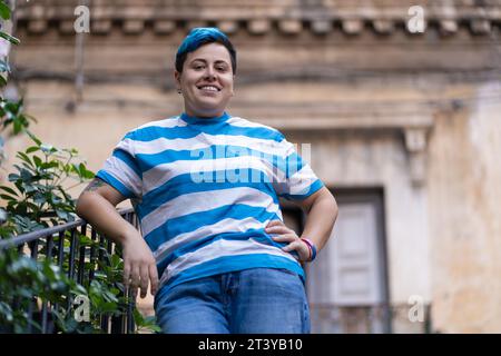 Giovane individuo gay con tatuaggio in ambiente urbano - persona con capelli blu che posa con fiducia da un balcone tra vecchi dettagli architettonici, ad esempio Foto Stock