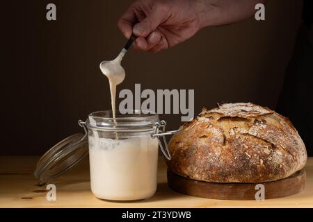 Le mani di un giovane che mostra com'è la pasta madre in un vaso di vetro. Con un pane crudo a pasta madre accanto a lui. Foto Stock