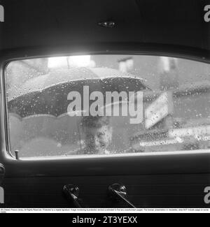 Negli anni '1950 E' un giorno piovoso nel centro di Stoccolma, l'anno è il 1954 e l'estate in cui la pioggia non sembrava mai volersi fermare. Una donna sta sotto un ombrello sotto la pioggia e sembra ancora felice e sorridente. È chiaro che il tempo è davvero piovoso, poiché viene fotografata dall'interno di un'auto attraverso il finestrino coperto da gocce di pioggia. Svezia 1954. Kristoffersson rif. BP92-2 Foto Stock