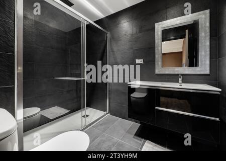 Il bagno è interamente piastrellato con ardesia nera, mobili di legno nero lucido e schermo in vetro Foto Stock