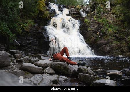 una giovane e bella turista seduta sullo sfondo di una cascata circondata da una foresta Foto Stock