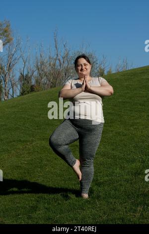 la donna di mezza età in sovrappeso pratica yoga all'aperto a piedi nudi, facendo esercizi di equilibrio su una gamba, posa sull'albero. Fitness, allenamento all'aperto, yoga, pesi l Foto Stock