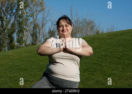 la donna di mezza età in sovrappeso pratica yoga all'aperto a piedi nudi, facendo esercizi di equilibrio su una gamba, posa sull'albero. Fitness, allenamento all'aperto, yoga, pesi l Foto Stock