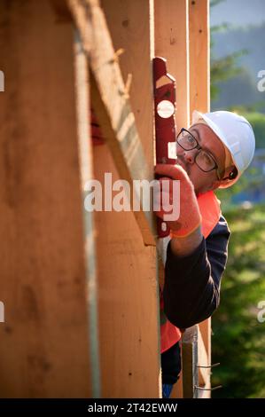 Carpentiere che costruisce una casa a telaio in legno vicino alla foresta. L'uomo barbuto, indossando occhiali, ispeziona le pareti per la livellatezza usando la livella a bolla d'aria. È vestito con casco protettivo e giubbotto arancione. Foto Stock