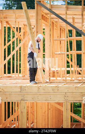 Carpenter costruisce una casa con struttura in legno. L'uomo tiene in mano un grande fascio mentre indossa abiti da lavoro e casco. Idea di costruzione moderna ed ecologica. Foto Stock