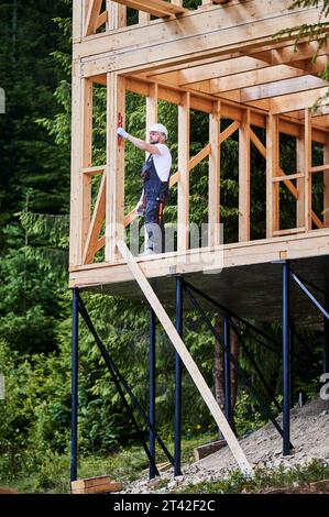 Carpenter costruisce una casa a due piani con struttura in legno nelle immediate vicinanze dei boschi. L'uomo barbuto, vestito con tuta e casco protettivo, valuta il livellamento del muro con l'aiuto di una livella. Foto Stock