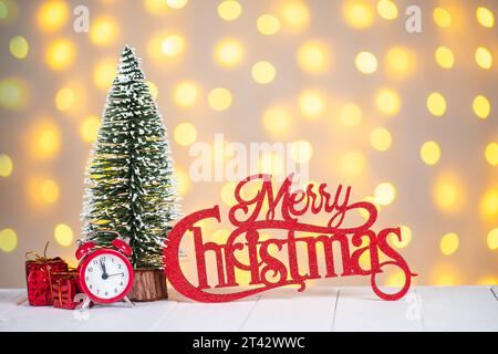 Albero decorativo di Natale, sveglia e scritta in legno rosso buon Natale su sfondo bokeh chiaro sfocato con spazio copia. Visualizzazione delle lancette dell'orologio Foto Stock