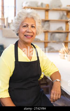 Felice ceramista birazziale con capelli grigi, seduta e sorridente nello studio di ceramica Foto Stock