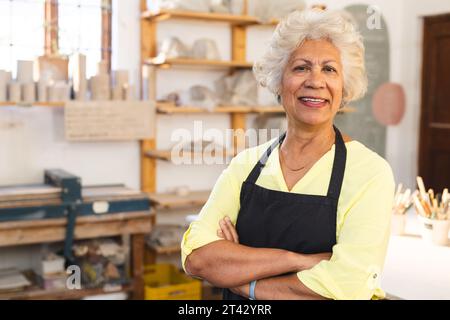 Felice ceramista birazziale anziana con capelli grigi, in piedi e sorridendo nello studio di ceramica Foto Stock