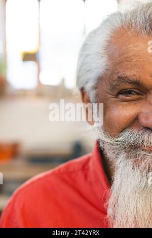 Mezza faccia di un uomo anziano birazziale felice con la barba lunga, sorridendo in uno studio di ceramica Foto Stock