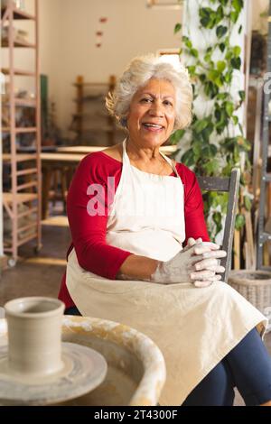 Felice ceramista birazziale anziana con capelli grigi seduta accanto alla ruota di potter nello studio di ceramica Foto Stock