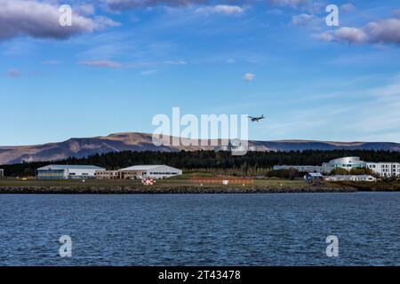 Reykjavik, Islanda - 25 settembre 2023: Paesaggio con aeroporto nazionale di Reykjavik e edificio Perlan. Un piccolo aereo in aria, una baia d'acqua di fronte. Foto Stock