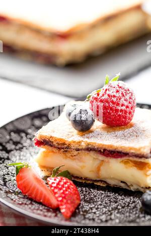 Un pezzo di torta Napoleone con semi di papavero e ciliegie, decorato con fragole, mirtilli su sfondo chiaro. Primo piano Foto Stock