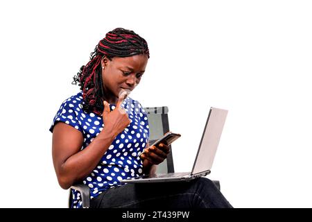 una bella donna d'affari con trecce seduta sulla sedia a casa che legge messaggi sul cellulare con un look attento mentre tiene il computer portatile sulle cosce. Foto Stock